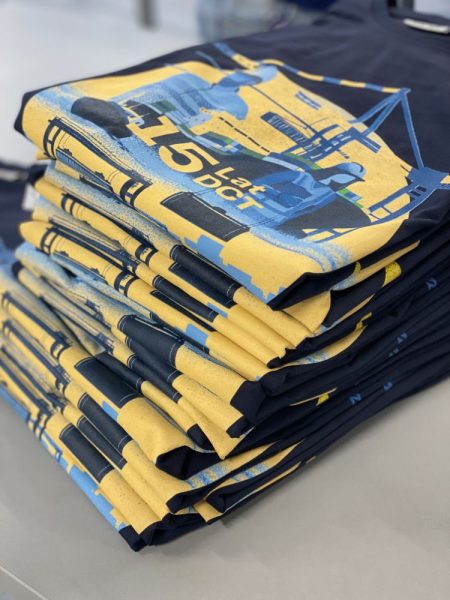 Stos granatowych t-shirtów z żółto-niebieskim nadrukiem, motywem portowym z napisem 15 lat DCT