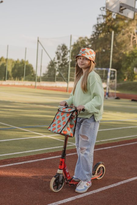 Uśmiechnięta dziewczynka na hulajnodze z lunchboxem i w kapeluszu z printem truskawkowym